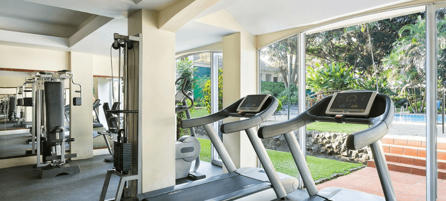 Bright and modern fitness center with treadmills, weight machines, mirrored walls, and large floor-to-ceiling windows overlooking a lush garden and outdoor pool area.