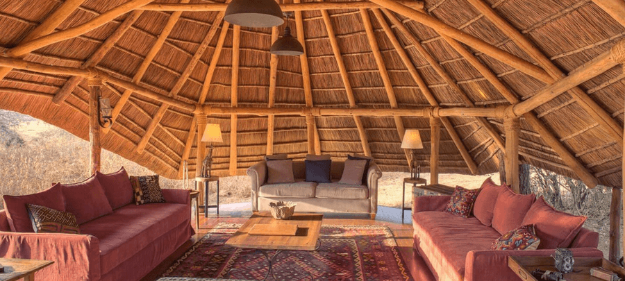 A cozy open-air lounge with thatched roofing, wooden beams, plush sofas, and patterned rugs offers a rustic retreat overlooking the surrounding bush landscape.