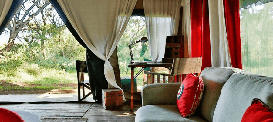A cozy safari tent lounge area with soft natural light filtering through large mesh windows and draped white curtains. The space features a cushioned sofa with bright red accent p…