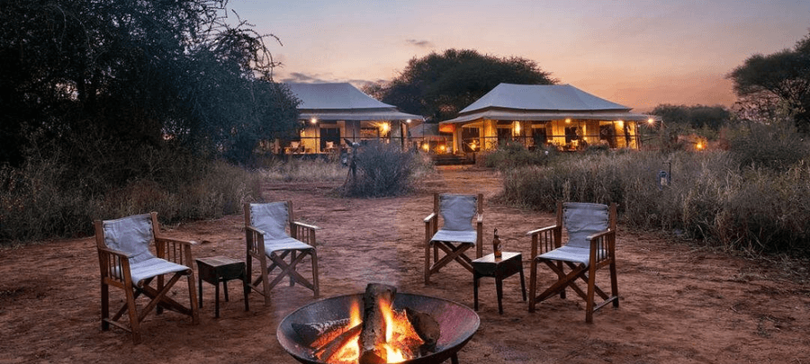 An outdoor safari campfire setup sits at the center of a rustic clearing at dusk, surrounded by wooden canvas chairs arranged for guests to relax and socialize. In the background,…