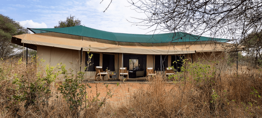 A spacious luxury safari tent sits surrounded by dry bushland and scattered trees, blending naturally into the wilderness. The tent features a shaded veranda with wooden rocking c…