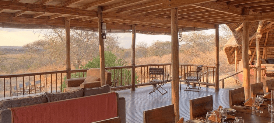 An open-air wooden lodge deck with lounge seating and a dining area overlooks the dry savannah landscape in soft golden light.