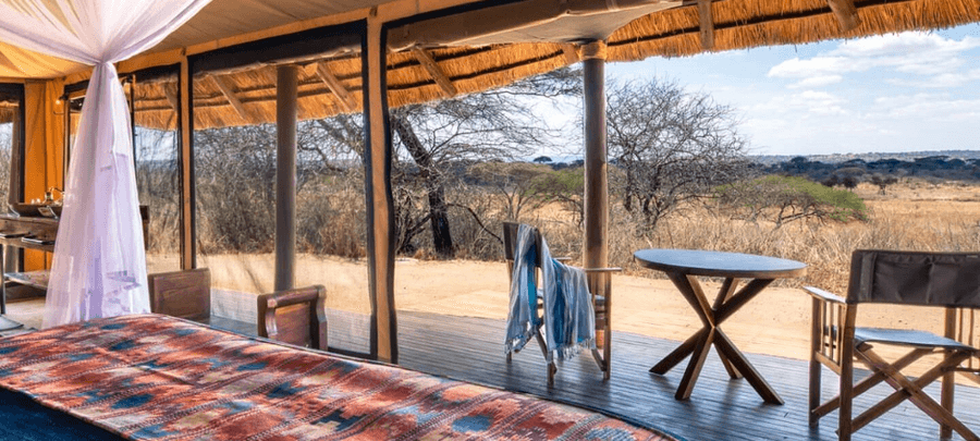 The interior of a luxury safari tent features a bed with a patterned blanket, open mesh walls, and a private deck with chairs overlooking the dry savannah landscape.