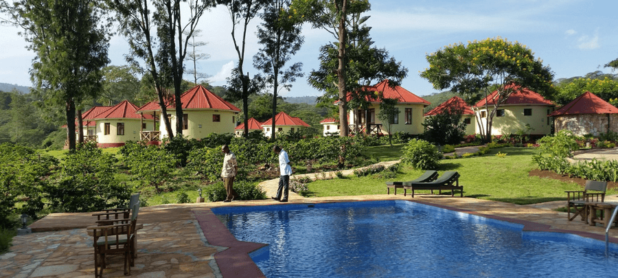 A peaceful outdoor resort scene featuring a swimming pool in the foreground, surrounded by lounge chairs and stone paving. In the background, several round cottages with red roofs…