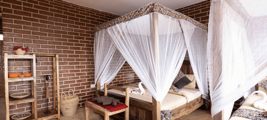 A rustic bedroom with exposed brick walls and wooden furniture, featuring canopy beds draped in white mosquito netting. Decorative woven baskets, handcrafted decor, and soft natur…
