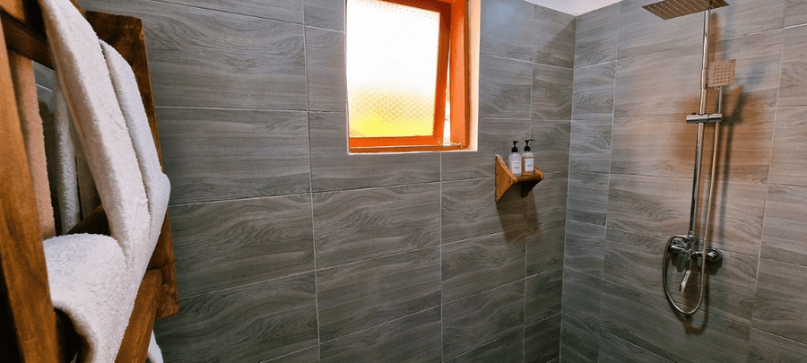 A modern bathroom shower area with gray tiled walls, a rainfall showerhead, and a small frosted window letting in warm light. White towels hang on a wooden rack beside neatly plac…