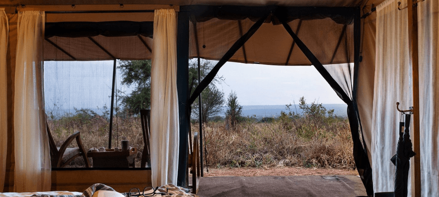 The open entrance of a luxury safari tent frames peaceful views of dry grassland and distant wilderness. Soft curtains, screened walls, and outdoor seating create a calm indoor-ou…