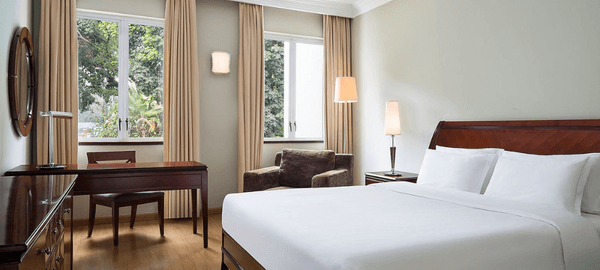 Modern and cozy hotel room with a large white bed, wooden furniture, soft lighting, beige curtains, and large windows overlooking lush greenery.