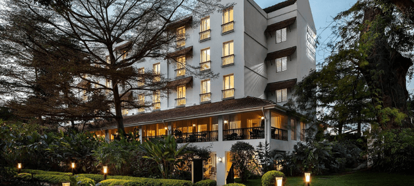 Elegant multi-story hotel surrounded by lush greenery, with warmly lit balconies and outdoor seating areas glowing at dusk.