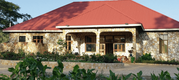 A large stone-clad lodge building with a bright red metal roof, surrounded by landscaped gardens and greenery. The entrance features arched walkways, large windows, and a paved co…