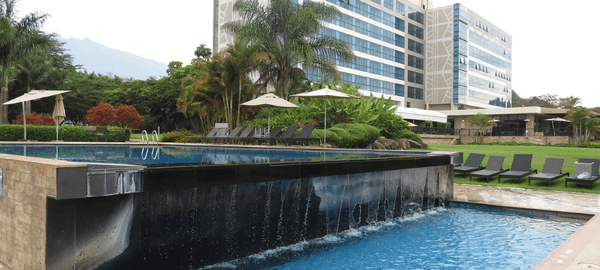 Luxury hotel pool area with cascading water features, sun loungers, tropical landscaping, and a modern multi-story hotel building in the background.