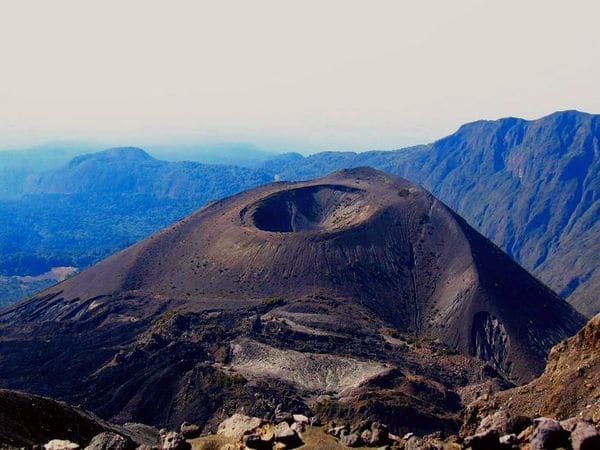 An aerial view of Mount Meru featuring its dramatic volcanic crater, rugged ash-covered slopes, and surrounding mountain landscape beneath a hazy sky.