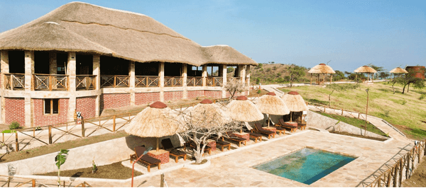 A spacious safari-style lodge with a large thatched roof overlooks a pool terrace lined with cushioned loungers under straw umbrellas. The property sits on gently rolling grassy h…