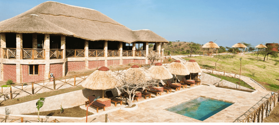 A spacious safari-style lodge with a large thatched roof overlooks a pool terrace lined with cushioned loungers under straw umbrellas. The property sits on gently rolling grassy h…