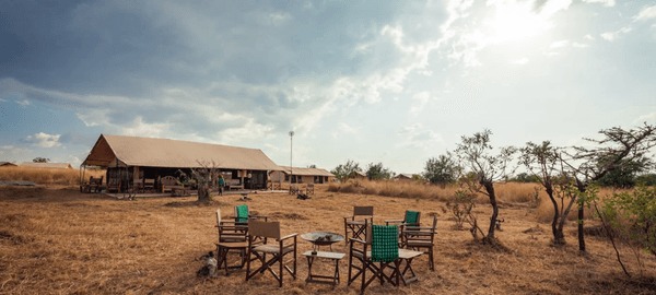 GNU Camp safari tented lodge set in open savannah, with outdoor chairs around a fire pit and main tent under a partly cloudy sky.