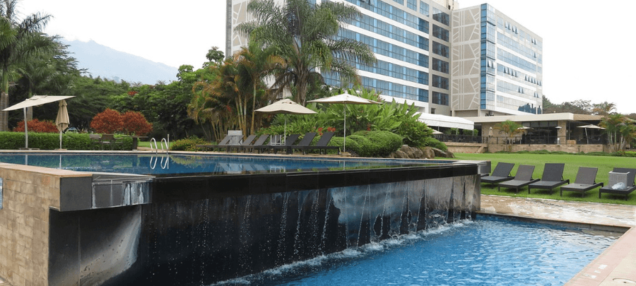 Luxury hotel pool area with cascading water features, sun loungers, tropical landscaping, and a modern multi-story hotel building in the background.