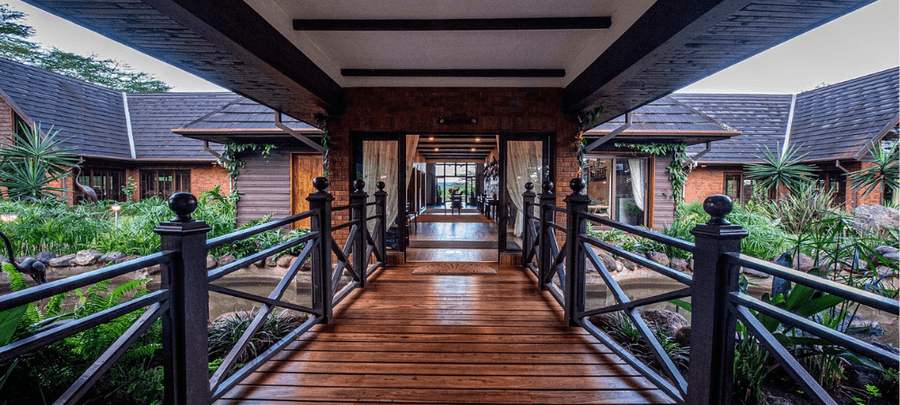 Elegant wooden walkway leading through a lush tropical courtyard to a luxury lodge entrance, surrounded by landscaped gardens, water features, and modern rustic architecture.