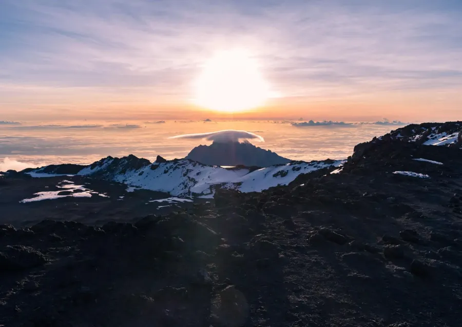 Sunrise above snowy volcanic terrain, with a distant mountain peak rising through clouds beneath a glowing sky.